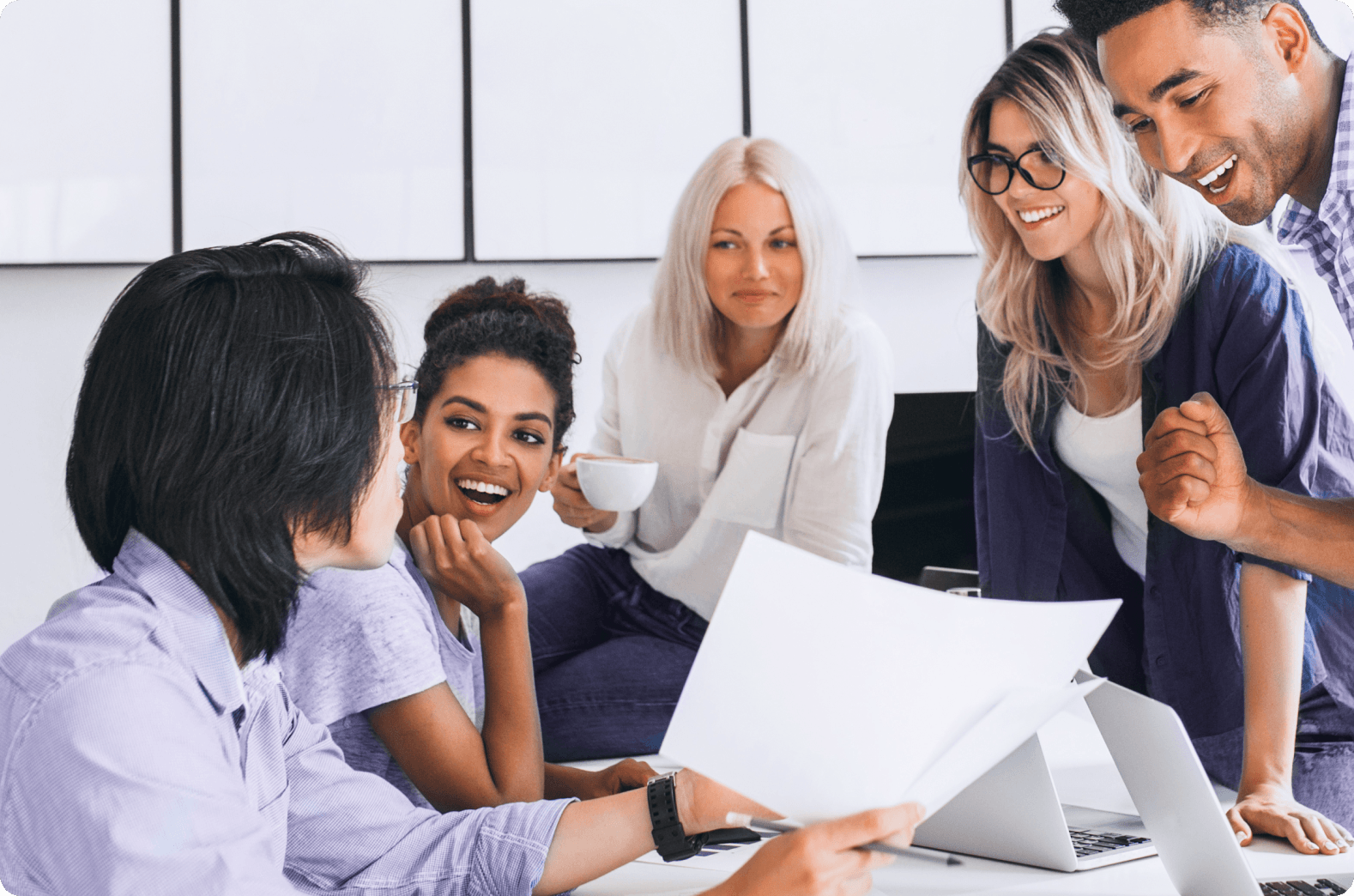 A diverse group of five smiling professionals collaborating around a table with laptops and documents, depicting teamwork and a positive work environment at an SEO agency.